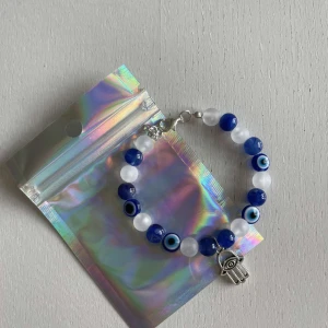 Egengjort pärlarmband  - Egengjort pärlarmband med blåa och vita pärlor. Armbandet har en berlock som avbildar en hand med ett öga inuti. 