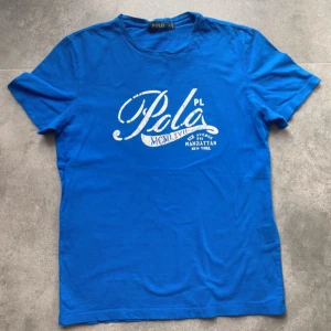 Blå t-shirt från Polo - vi låter ai skriva om denna grymma sommartisha denna gång riktigt laidback om du frågar mig. En blå t-shirt från Polo med ett tryck i vitt på framsidan som inkluderar texten 'Polo PL' och 'Manhattan New York'. T-shirten har korta ärmar och en rund halsringning.