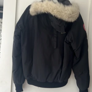 Svart jacka från Canada Goose - En svart jacka från Canada Goose med en pälsfodrad huva. Jackan har en dragkedja och knappar framtill samt flera fickor. Märkets logotyp är synlig på ärmen.