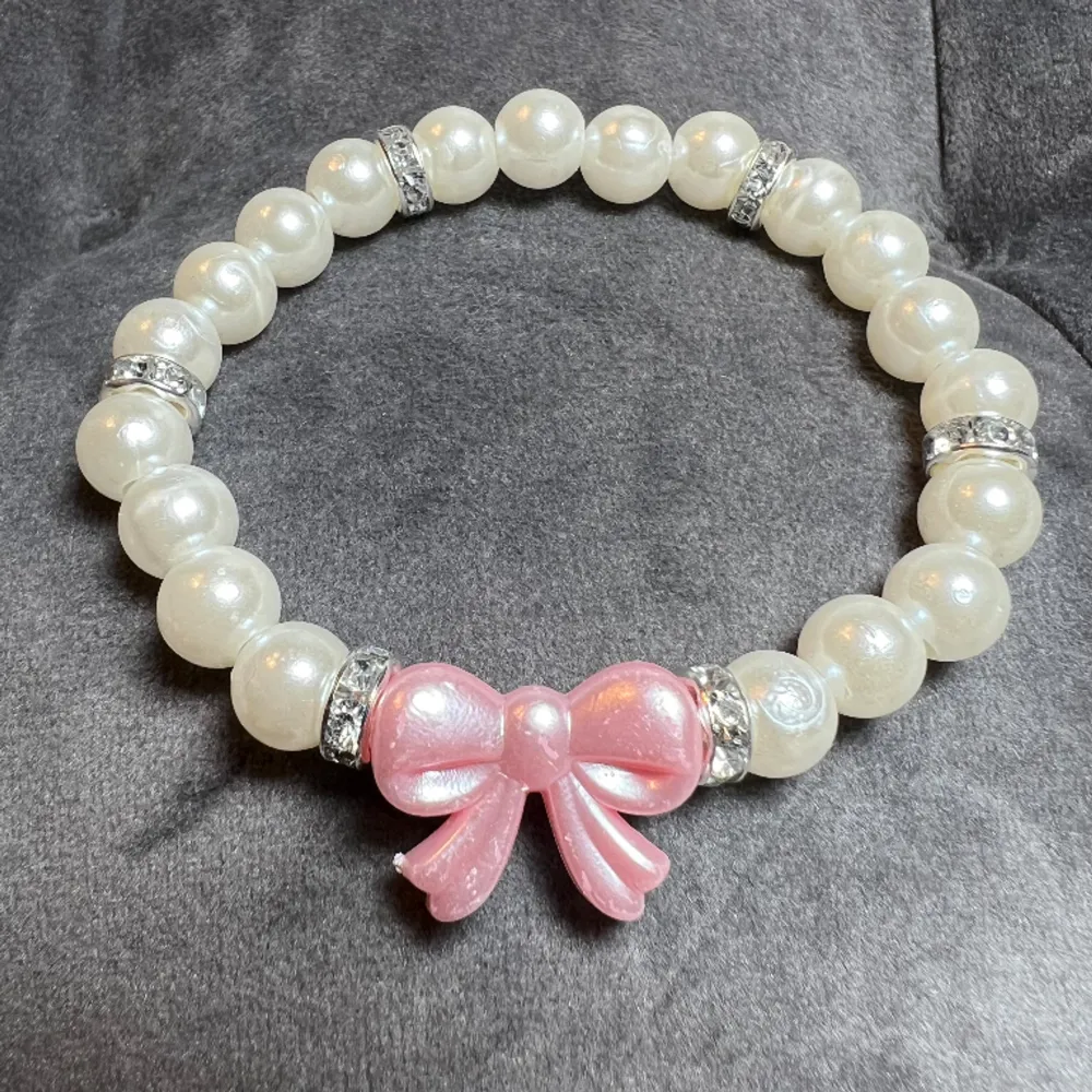 Egentillverkat pärlarmband med vita pärlor, blingiga mellanplattor och en söt rosa rosett.🎀Är gjort med gummitråd och är ca 16 cm långt.. Asusteet.