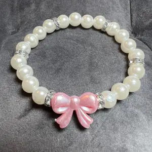 Egentillverkat pärlarmband med vita pärlor, blingiga mellanplattor och en söt rosa rosett.🎀Är gjort med gummitråd och är ca 16 cm långt.