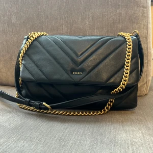 DKNY väska  - Säljer min DKNY väska i svart och guld. Den är använd med är fortfarande i fint skick.  Reglerbart axelband.  Dustbag medföljer. 