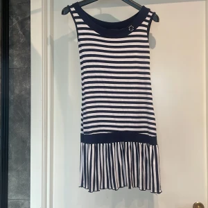 Randig klänning från Natural - En ärmlös randig klänning från märket Natural. Klänningen har horisontella ränder i marinblått och vitt, med en bredare marinblå rand vid midjan och en plisserad nederdel. Den har en rund halsringning och en dekorativ knapp vid halsen.