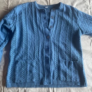 Blå stickad kofta - En blå stickad kofta med ett mönster i tyget. Koftan har långa ärmar, knappar framtill och två fickor på framsidan. Materialet består av 50% ull och 50% akryl. tyvärr saknas en knapp 