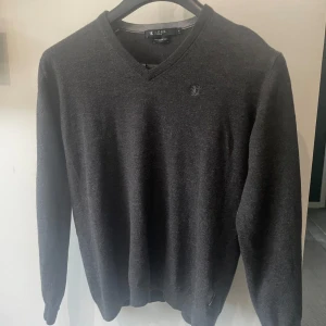 Mörkgrå tröja från Tiger of Sweden - En stilren och bekväm mörkgrå tröja från Tiger of Sweden. Tröjan har en v-ringad hals och är tillverkad av mjukt merinoull material. Den sitter dom S men är storlek L
