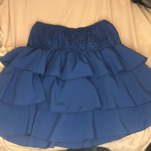 Blå volangkjol - En blå kortkjol med volanger och elastisk midja. Kjolen har flera lager av volanger som ger en lekfull och feminin look.den köptes i Italien 