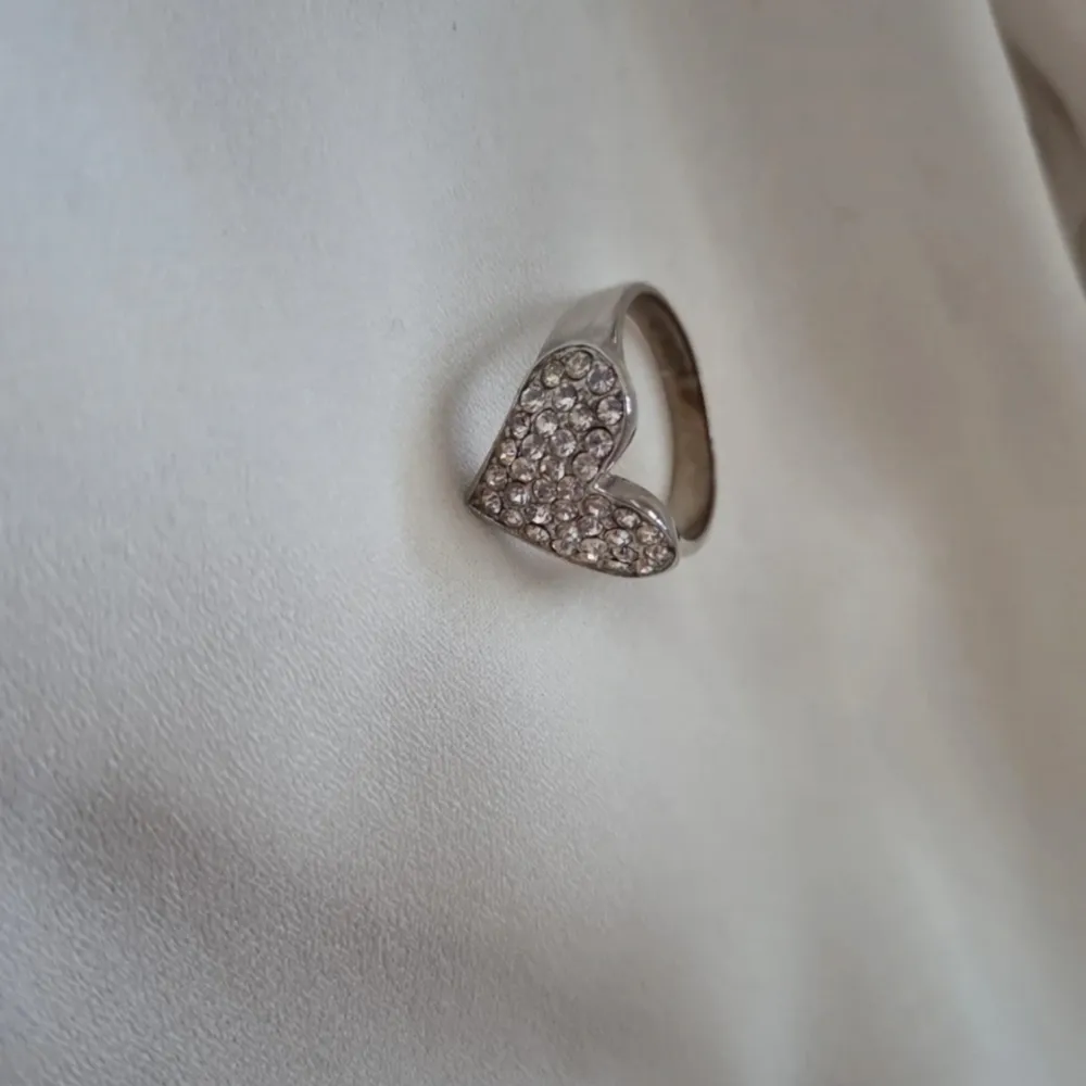 En silvrig ring med ett stort hjärta täckt av diamanter på. Dessa är troligtvis fake och ringen tror jag är i någon metall.. Accessoarer.