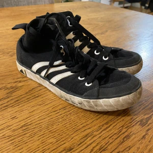 Adidas tygskor - Svarta adidas tygskor med vita ränder. Den vita sulan har gulat en aning och har diverse märken (syns på bilderna), men resten av skon är i fint skick.