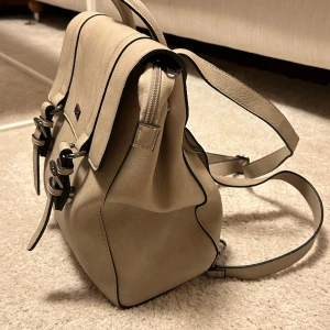 Väska Don Donna - Beige väska från Don Donna.  Mycket fint skick!  Skinnimitation Går att ha både som ryggsäck, over shoulder och cross body.  Magnetspännen på locket.  Mått: 25x30x12