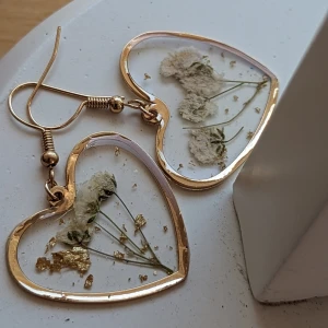 Hängande Örhängen - Designade med en detaljrik blomma i form av ett hjärta, kommer de att vara ett fantastiskt tillskott till din smyckesamling.  Design: Blomma formad som ett hjärta Stil: Hängande örhängen Skick: Mycket fint skick, nästan som nya Material ospecificerat
