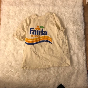 Fanta T-shirt  - En vit T-shirt med en blå text där det står fanta och en orange text där det står orange över är de apelsiner med ett grönt blad och under är de 1 ljud orange ett orange och ett blåt