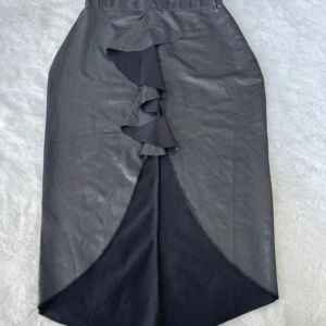 Svart kjol med volangdetalj - En elegant svart kjol med volangdetalj framtill. Kjolen har en asymmetrisk design och är tillverkad i ett glansigt material.