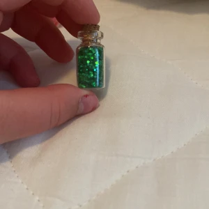 Grönt glitter i liten glasflaska - En liten glasflaska fylld med grönt glitter. Flaskan har en kork som förslutning och är perfekt för dekorativa ändamål eller pyssel.