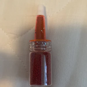 Röd ögonskugga i flaska - En flaska med röd ögonskugga i form av små pärlor. Flaskan har en genomskinlig kropp och en orange topp med en vit applikator.