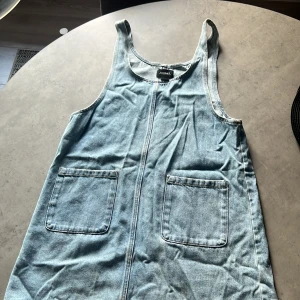 Jeansklänning från Monki - En ärmlös jeansklänning från Monki i storlek S. Klänningen har två framfickor. Den är tillverkad i ett ljusblått jeanstyg.