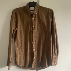 Brun skjorta från Lindbergh - En brun långärmad skjorta från Lindbergh i slim fit. Skjortan är tillverkad av ekologisk bomull och har knappar framtill samt vid ärmsluten. storlek M. bra skick