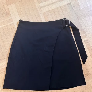 Svart kjol från Monki - En svart kjol från Monki med omlott-design och ett bälte med spänne. Kjolen är kort och har en stilren look. Har tagits väl hand om. Storleken går mot 32/34. 