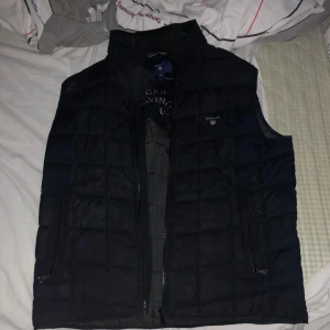 Svart dunväst från GANT - En svart dunväst från GANT med dragkedja framtill. Västen har en quiltad design och är märkt med GANT-logotypen på bröstet. Den är i storlek M och är lämplig för höst- och vintersäsonger.