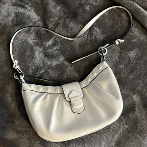 Vit handväska - Vit handväska från river island. Den har lite svara detaljer och bandet är justerbart så man kan ha den hur lång man vill. Den har ett magnetiskt lås. Köpt på second hand.