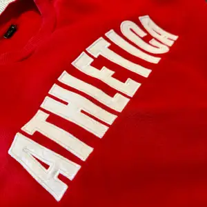 Röd sweatshirt med texten ”Athletica” på framsidan. Bra skick.