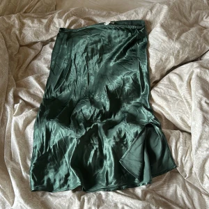Mörkgrön sidenklänning - Säljer en elegant mörkgrön kjol i siden. kjolen går nedanför knänen och har en slits på sidan som ger en snygg detalj. Perfekt för fest eller andra speciella tillfällen. Den är i nyskick och har en vacker glans som gör att den sticker ut. Passar perfekt för höst och vinter. Kjolen går att stryka fast ut och in i  lägre temperaturer (inte strykt på bild)