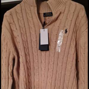 Säljer en snygg beige stickad tröja från Polo Ralph Lauren. Tröjan har en dragkedja vid halsen och är dekorerad med det klassiska Polo Ralph Lauren-loggan på bröstet. Perfekt för höst och vinter, den är både stilren och varm. Passar bra till både vardag och fest!