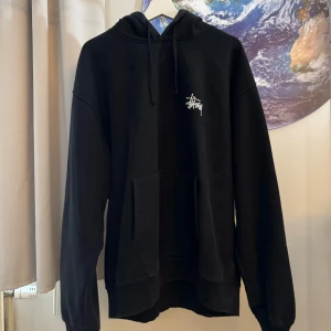 Svart hoodie från Stüssy - Säljer en svart hoodie från Stüssy i storlek XL. Den är superbekväm och perfekt för höst och vinter. Hoodien har en stor ficka på framsidan och Stüssy-loggan tryckt både fram och bak. Den är i bra skick och redo för en ny ägare!