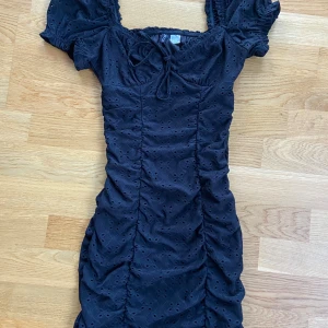 Svart kortklänning från H&M - Säljer en superfin svart kortklänning från H&M i storlek XXS. Klänningen har korta puffärmar och en söt knytning framtill. Den är tillverkad i ett mönstrat tyg med små hål och har en tight passform som framhäver figuren. Perfekt för fest eller en utekväll! 💃