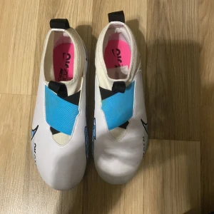 Nike Air Zoom fotbollsskor - Säljer ett par Nike Air Zoom fotbollsskor i nyskick. Skorna är vita med blå och rosa detaljer, inklusive en blå sula med rosa dobbar. De har en modern design med en elastisk blå rem över vristen och en dragflik på hälen för enkel påtagning. Perfekta för fotbollsträning och matcher.
