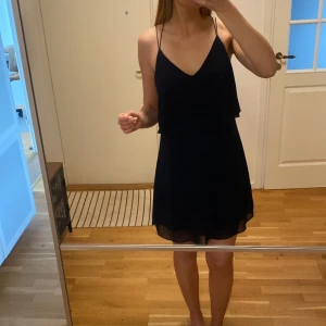 Mörkblå klänning med tunna axelband - Mörkblå klänning med tunna axelband och korsade band i ryggen. Klänningen har en lös passform och faller fint. Den har en layer-effekt upptill som ger intryck av ett markera midjan lite. Djup rygg. 