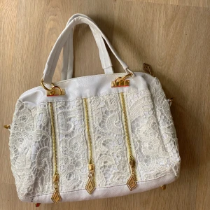 Vit handväska med spetsdetaljer och gulddragkedjor - Säljer en superfin vit handväska med spetsdetaljer och gulddragkedjor. Väskan har två handtag och en dragkedja upptill. Insidan är fodrad med ett blommigt mönster och har flera fack för förvaring. Perfekt för både vardag och fest! 👜✨ 20*30 cm