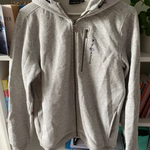 Grå hoodie från Sail Racing - Säljer en grå hoodie från Sail Racing i storlek 176. Den har en dragkedja framtill och en bröstficka med dragkedja. Hoodien är långärmad och har en bekväm passform. Perfekt för vardagsbruk eller som ett extra lager under kyligare dagar.