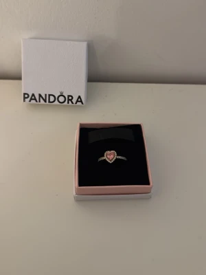 Pandora ring med rosa hjärta - Söt ring från Pandora i guldton med ett rosa hjärta i mitten och små gnistrande stenar längs bandet. Kommer i original ask. Perfekt för dig som gillar romantiska detaljer och vill ha något extra fint på fingret.