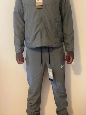 Grått träningsset från Nike - Säljer ett grått träningsset från Nike med jacka och byxor. Setet har en sportig look med dragkedja på jackan, resår i midjan och muddar vid bensluten. Perfekt för träning eller chill. Materialet är lätt och andas bra.