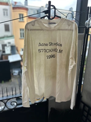 Vit långärmad tröja Acne Studios - Vit långärmad topp från Acne Studios med tryck 'Acne Studios STOCKHOLM 1996' på bröstet. Tillverkad i ett tunt och mjukt material med avslappnad passform och rund halsringning. Perfekt för en clean och stilren look.
