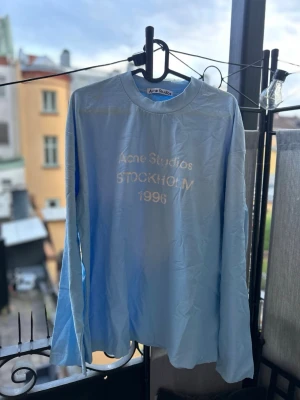 Ljusblå långärmad topp Acne Studios - Ljusblå långärmad topp från Acne Studios med tryck 'STOCKHOLM 1996' framtill. Tillverkad i mjukt material och har en klassisk rund halsringning. Perfekt för en avslappnad och stilren look.