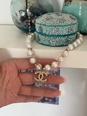 Pärlhalsband med glittrig CC-berlock - Snyggt halsband med stora vita pärlor och guldfärgade detaljer. Halsbandet har en glittrig CC-berlock täckt av små stenar som ger extra bling. Perfekt accessoar för att lyfta din outfit och ge en lyxig känsla.