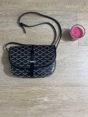 Svart axelväska med mönster - Snygg svart axelväska från Goyard med klassiskt vitt mönster och justerbar axelrem. Väskan har ett lock med spänne och detaljer i svart läder. Perfekt för dig som gillar stilrena accessoarer med lyxig känsla.