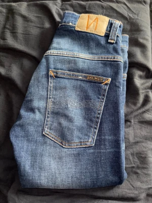 Blå jeans från Nudie Jeans - Säljer ett par klassiska blå jeans från Nudie Jeans med snygga slitningar och kontrastsömmar. Jeansen har fem fickor, normal midja och raka ben. Tillverkade i mjukt denimtyg som ger en skön passform. Perfekta för en avslappnad stil.