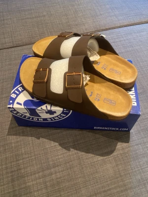 Birkenstock - Säljer ett par klassiska Birkenstock sandaler i brunt med två breda remmar och justerbara metallspännen. Fotbädden är mjuk och i beige mocka för extra komfort. Perfekta för dig som vill ha både stil och bekvämlighet.