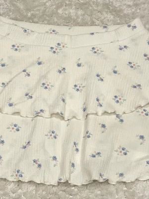 Vit blommig volangkjol - Söt kort kjol i vit färg med små blå och rosa blommor. Kjolen har volangdetaljer och är gjord i ett lätt, strukturerat material som känns luftigt. Perfekt för varma dagar och ger en romantisk vibe till din outfit.