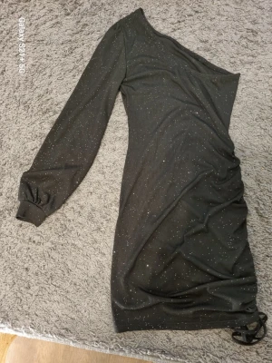 Svart glittrig oneshoulder klänning - Snygg svart kortklänning med glittriga detaljer och oneshoulder-design. Klänningen har lång ärm på ena sidan och rynkade detaljer i sidan. Perfekt för dig som vill sticka ut med en trendig look.