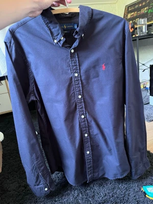 Mörkblå skjorta från Ralph Lauren - Klassisk mörkblå skjorta från Ralph Lauren med knappslå och vit knappdetalj. Skjortan har lång ärm och den ikoniska röda logotypen broderad på bröstet. Tillverkad i bomull och har en tidlös, normal passform.