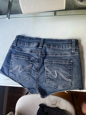 Mörkblå jeansshorts med rå kant - Säljer ett par mörkblå jeansshorts med rå, fransig kant och dekorativa bakfickor med vit söm. Shortsen har hög midja, bälteshällor och dubbla knappar framtill. Perfekta för sommaren och ger en avslappnad vibe. Vet inte riktig vad dom kosade nypris