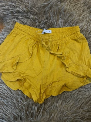 Gula linneshorts med volang från Sister - Snygga gula shorts från Sister i linne med volangdetaljer längs sidorna och knytband i midjan. De har en högmidjad passform och elastisk midja för extra komfort. Perfekta för varma dagar och ger en fräsch sommarkänsla.