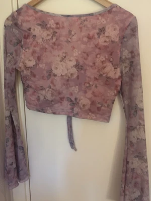 Blommig mesh topp med snörning - Säljer en croppad, långärmad topp i mesh med blommigt mönster i rosa och lila toner. Toppen har snörning framtill och lätt utsvängda ärmar. Perfekt för dig som gillar romantisk och trendig stil.