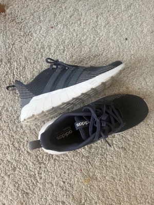 Adidas grå sneakers med vita sulor - Snygga sneakers från Adidas i mörkgrått med klassiska tre ränder på sidan. Funkar bra för att löpa med såsom vardagsakiveter. Bara höra av sig vid funderingar!