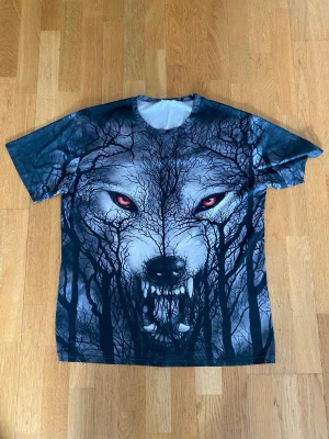 T-shirt med vargtryck och röda ögon - Unik t-shirt med ett stort tryck av en varg med intensiva röda ögon och trädgrenar som formar ansiktet. T-shirten har korta ärmar och är i mörka toner av svart, grått och blått. Perfekt för dig som gillar djurmotiv och vill sticka ut.