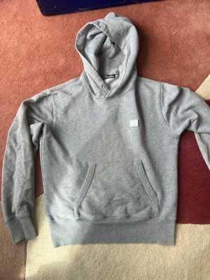 Grå hoodie från Acne Studios - Säljer en stilren grå hoodie från Acne Studios med deras ikoniska ansiktslogga på bröstet. Hoodien har huva med dragsko, känguruficka och ribbade muddar. Tillverkad i mjuk bomullsblandning som är perfekt för chill dagar.