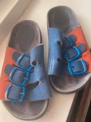 Blå och röda sandaler med spännen(barn) - Säljer ett par sandaler i blått och orange med tre justerbara spännen i blå plast på varje sandal. Sulan är grå och har en formad fotbädd för extra komfort. Perfekta för sommardagar eller att glida runt hemma.
